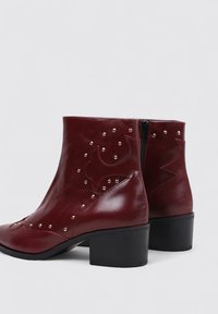 Bottines en cuir rouge avec un bout pointu, un talon carré, des clous décoratifs et des coutures, équipées d'une fermeture éclair sur le côté.
