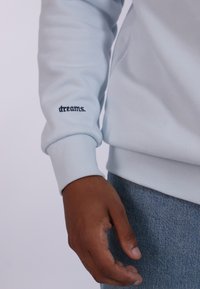 Felpa azzurra con polsini a coste, con la scritta ricamata "dreams." sulla manica. Tessuto morbido, abbinata a jeans in denim.