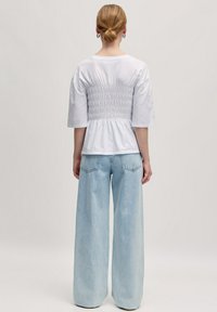 Witte T-shirt met een gerimpelde achterkant, gecombineerd met losse lichtblauwe jeans. De outfit heeft een relaxte pasvorm en een eenvoudig ontwerp.