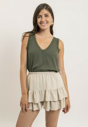 Femme souriante portant un haut sans manches vert olive et une jupe beige à volants superposés, debout devant un fond clair uni.