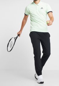 Polo shirt vert clair avec des accents et un logo noirs, associé à un pantalon de sport noir. La personne tient une balle de tennis et une raquette.