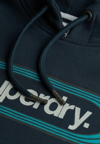 Granatowy hoodie z przednią kieszenią. Posiada turkusowe i brązowe poziome paski. Z przodu znajdują się sznurki z metalowymi końcówkami oraz detal z logo.