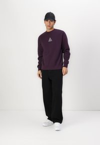 Violetinis džemperis su apvaliu kaklu ir baltu trikampio logotipu ant krūtinės, derinamas su juodomis plačiomis kelnėmis ir baltomis sportinėmis bateliais. Mūvimas juodas kepurė.