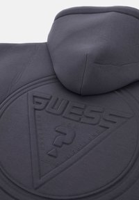 Sudadera con capucha de color gris oscuro que presenta un logo triangular en relieve con "GUESS" y otros detalles textuales. Textura suave con un tacto agradable.