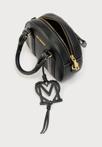 Μαύρη δερμάτινη τσάντα χειρός με στρογγυλεμένο σχέδιο, χρυσό φερμουάρ, λευκές ραφές και διακοσμητικό μενταγιόν λογότυπου.