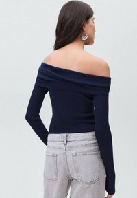 Námořnický svetr s odhalenými rameny a vroubkovaným vzorem, dlouhými rukávy, v kombinaci se světle šedými slim fit džíny. Hladká textura a jemný skládací detail na krku.