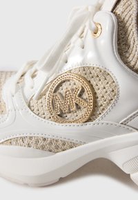 Közelkép egy bézs és fehér tornacipőről, texturált anyaggal, fehér fűzőkkel és arany MK logóval az oldalán.