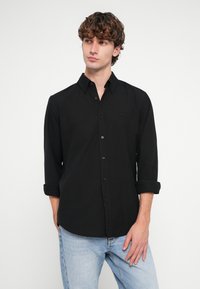 Μαύρο πουκάμισο με κουμπιά και γιακά, μακριά μανίκια τυλιγμένα στους αγκώνες και μια διακριτική λεπτομέρεια λογότυπου. Φορεμένο με ανοιχτόχρωμα μπλε τζιν.