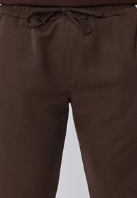 Gros plan sur un pantalon marron avec une taille élastique et un cordon de serrage noué à l'avant, montrant la texture lisse du tissu.