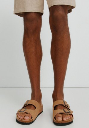 Benen met beige korte broek en lichtbruine suède sandalen met dubbele bandjes en zwarte gespen op een effen witte achtergrond.