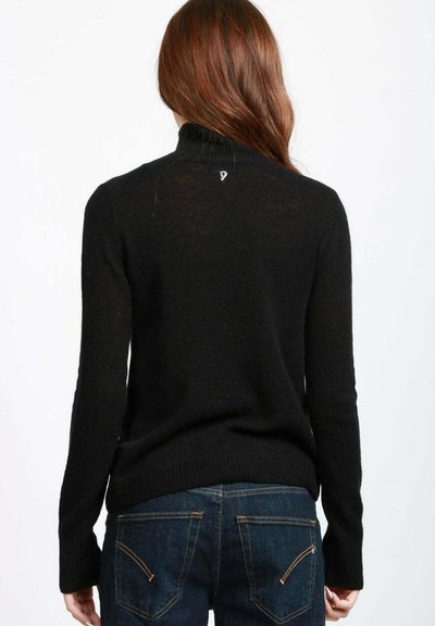Donna con lunghi capelli castani che indossa un maglione nero a collo alto e jeans blu, vista di spalle su uno sfondo neutro.