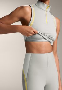 Light grey athletic tank top with a high collar, yellow accent stripes, and a zip closure, paired with matching high-waisted leggings.