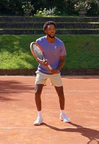 Tennis spiller i en lavendelfarvet skjorte og beige shorts, som holder en orange og grå tennisracket på en grusbane med grønne græs i baggrunden.