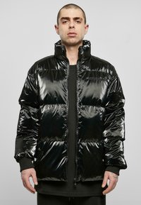 Doudoune noire avec une finition brillante, dotée d'un col montant, d'une fermeture éclair et de sections piquées. Portée par-dessus une chemise noire.