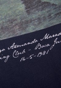 Donkere stof met een vervaagd afdruk van een voetballer, met witte handgeschreven tekst die "Diego Armando Maradona" en een datum vermeldt.