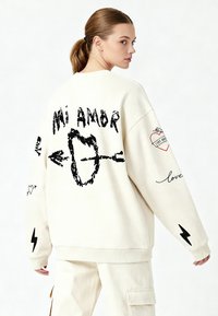 Felpa oversize color crema con grafiche nere, tra cui "Mi Amor", cuori e frecce sulla schiena e sulle maniche.