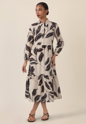 Frau trägt ein beiges Midikleid mit schwarzen abstrakten Blattmustern, langen Ärmeln, Kragen und geknöpftem Vorderteil, kombiniert mit schwarzen Riemchensandalen.