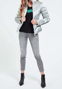 Sølvfarvet pufferjakke med quiltet mønster, sort tætsiddende skjorte med grønt grafisk motiv, slidte grå jeans og sorte støvler til anklen.