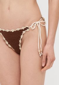Bruine gehaakte bikinibroek met een geschulpte crème rand. Beschikt over verstelbare strikken met houten kralen aan elke kant. Getextureerd ontwerp.