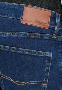 Niebieska tylna kieszeń dżinsów z denimu z przyszytą powyżej brązową skórzaną metką z logo Pepe Jeans London.