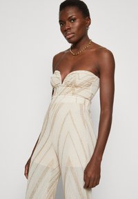 Trägerloser Jumpsuit in Beige mit goldenen Zickzackmustern, strukturiertem Stoff und figurbetonter Silhouette. Akzentuiert mit einem tiefen V-Ausschnitt und seitlichen Schlitzen.