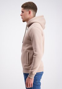 Smėlinis džemperis su priekiniais kišenėmis, margomis raišteliais ir raukinėtais rankogaliais. Dėvimas su mėlynomis džinsinėmis kelnėmis, rodomas iš šono ant balto fono.