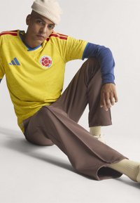 Jeune homme assis par terre portant un maillot de football jaune de Colombie, une chemise bleue à manches longues, un pantalon marron, des chaussettes beiges et un bonnet crème.