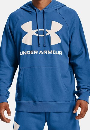 Mann trägt einen blauen Under Armour Kapuzenpullover und passende blaue Jogginghose, die Hände entspannt an den Seiten, sichtbar vom Hals bis zu den Oberschenkeln.