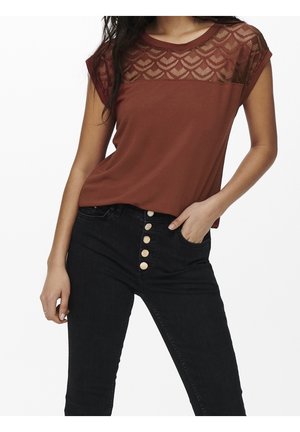 Vrouw draagt een roestkleurige top met korte mouwen en een patroon op het doorschijnende inzetstuk, gecombineerd met zwarte high-waist jeans met vijf gouden knopen.