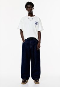 Hvid oversized T-shirt med trykt tekst på brystet, parret med mørkeblå brede denimbukser og brune chunky sko.