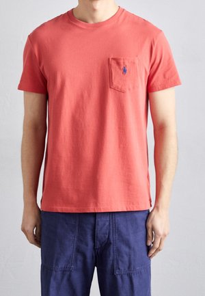 Man draagt een effen koraalrood T-shirt met korte mouwen en een borstzak, en blauwe casual broek, staand tegen een effen lichte achtergrond.