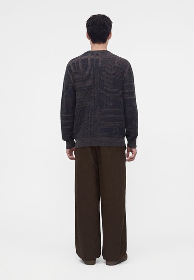 Homme debout de dos, vêtu d’un pull sombre à motifs et d’un pantalon large marron ample, sur un fond uni.