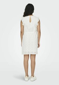 Weißes Kleid mit Lochmusterausschnitten, kurzen Rüschensäumen und einem Schlüsselaugen-Detail am Rücken. Verfügt über einen Gürtel für eine verstellbare Passform. Getragen mit weißen Turnschuhen.