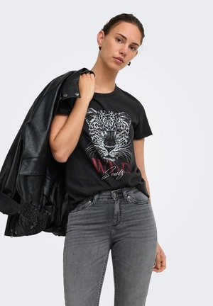 Jeune femme portant un t-shirt noir à imprimé léopard et un jean gris, tenant une veste en cuir noire sur son épaule devant un fond uni.