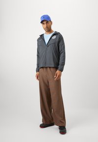 Chaqueta ligera gris con cremallera, camisa azul clara, pantalones ampulosos de cuadros marrones, y gorra azul, completado con zapatillas negras y rojas.