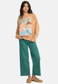 Bruine sweatshirt met grafische prints, losse fit; gecombineerd met teal wijd-pijpen broek; beige instap schoenen. Casual, ontspannen outfit.