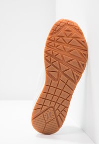 Weißer Sportschuh von unten betrachtet, mit einer strukturierten Gummisohle und einem dreieckigen Muster sowie einem seitlichen Logo-Detail.