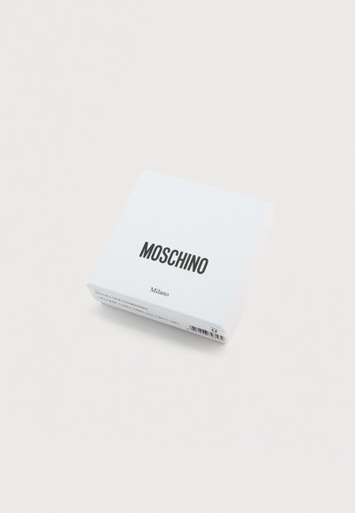 Λευκό κουτί με την επωνυμία Moschino Milano και γραμμωτό κώδικα στο πλάι, τοποθετημένο σε ανοιχτόχρωμη επιφάνεια.