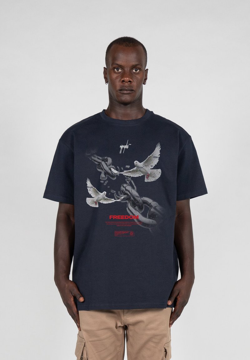 Donker marineblauw katoenen T-shirt met een afbeelding van brekende ketens en duiven, met daaronder in rode tekst het woord "VRIJHEID".