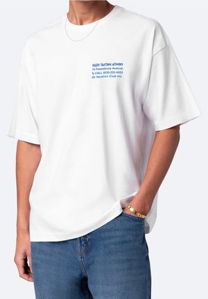 Osoba nosząca białą, oversize'ową koszulkę z niebieskim napisem „Ouzo Tasting Athens” i adresem na lewej piersi, w zestawieniu z niebieskimi dżinsami i złotą bransoletką.