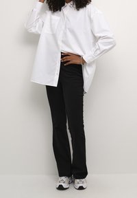 Chemise blanche à boutons avec une seule poche poitrine, associée à un pantalon noir ajusté et des baskets grises, sur un fond neutre.