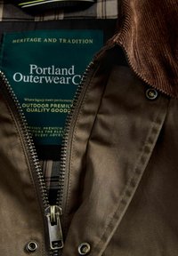 Zeleni kaput s vanjske strane s zatvaračem, ogroman ovratnik od kordura, podstavljene s kariranim uzorkom i oznakom "Portland Outerwear Co." s uputama za njegu.