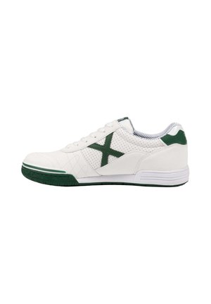 Zapatilla deportiva blanca de caña baja con detalle de cruz verde, acentos verdes en el talón y la suela, y paneles laterales perforados.