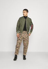 Bomber jacket verde oliva con polsini e fondo a costine, abbinata a pantaloni beige a camouflage. Collo alto nero sotto, stivaletti neri alla caviglia.