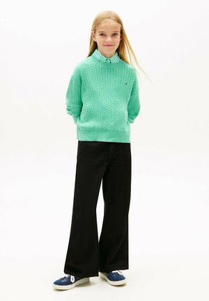 Ragazza con lunghi capelli biondi che indossa un maglione verde sopra una camicia a righe, pantaloni neri a gamba larga e scarpe da ginnastica blu, in piedi con le mani dietro la schiena.