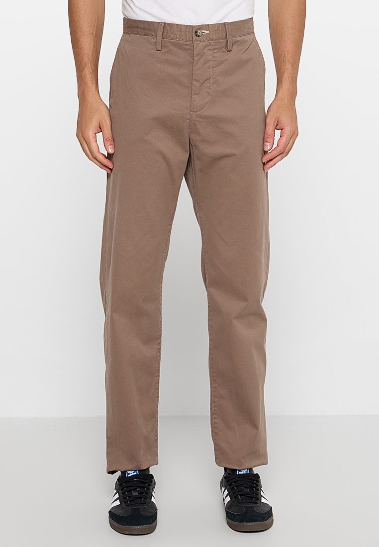 Braune Baumwollhosen mit entspannter Passform, ausgestattet mit einem Knopf- und Reißverschluss, Gürtelschlaufen und einer glatten Textur. Getragen mit schwarzen Sneakers.