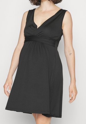 Femme en robe noire sans manches à col en V avec taille froncée et petit tatouage en forme de cœur sur l'avant-bras gauche, debout devant un fond uni.