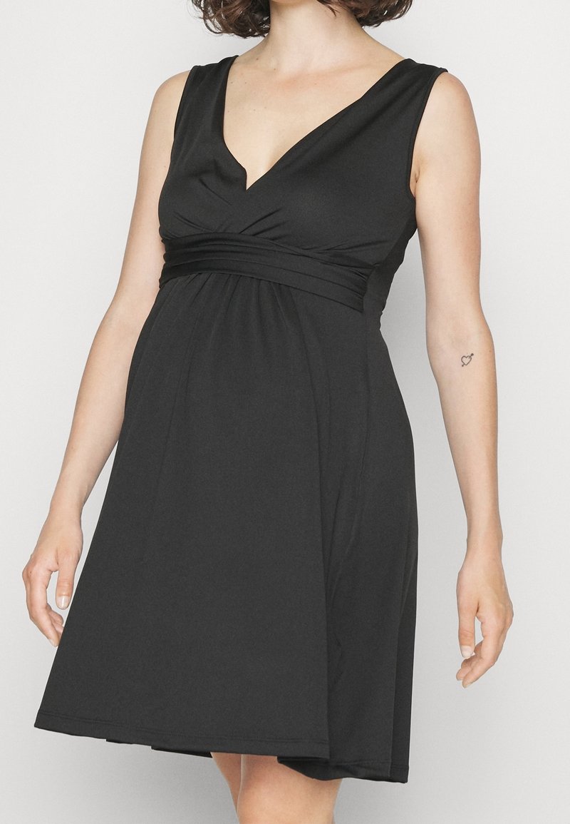 Femme en robe noire sans manches à col en V avec taille froncée et petit tatouage en forme de cœur sur l'avant-bras gauche, debout devant un fond uni.