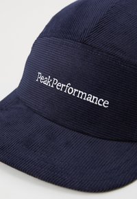 Tumesinine korduroy müts, millel on lame ääre, esipaneelil valge tikandtekst "PeakPerformance". Tekstuuriga pinnad.