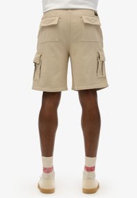 Pantaloni cargo beige con molte tasche laterali, vestibilità dritta e dettagli impreziositi da cuciture, realizzati in tessuto morbido, abbinati a scarpe beige e calzini a fantasia.